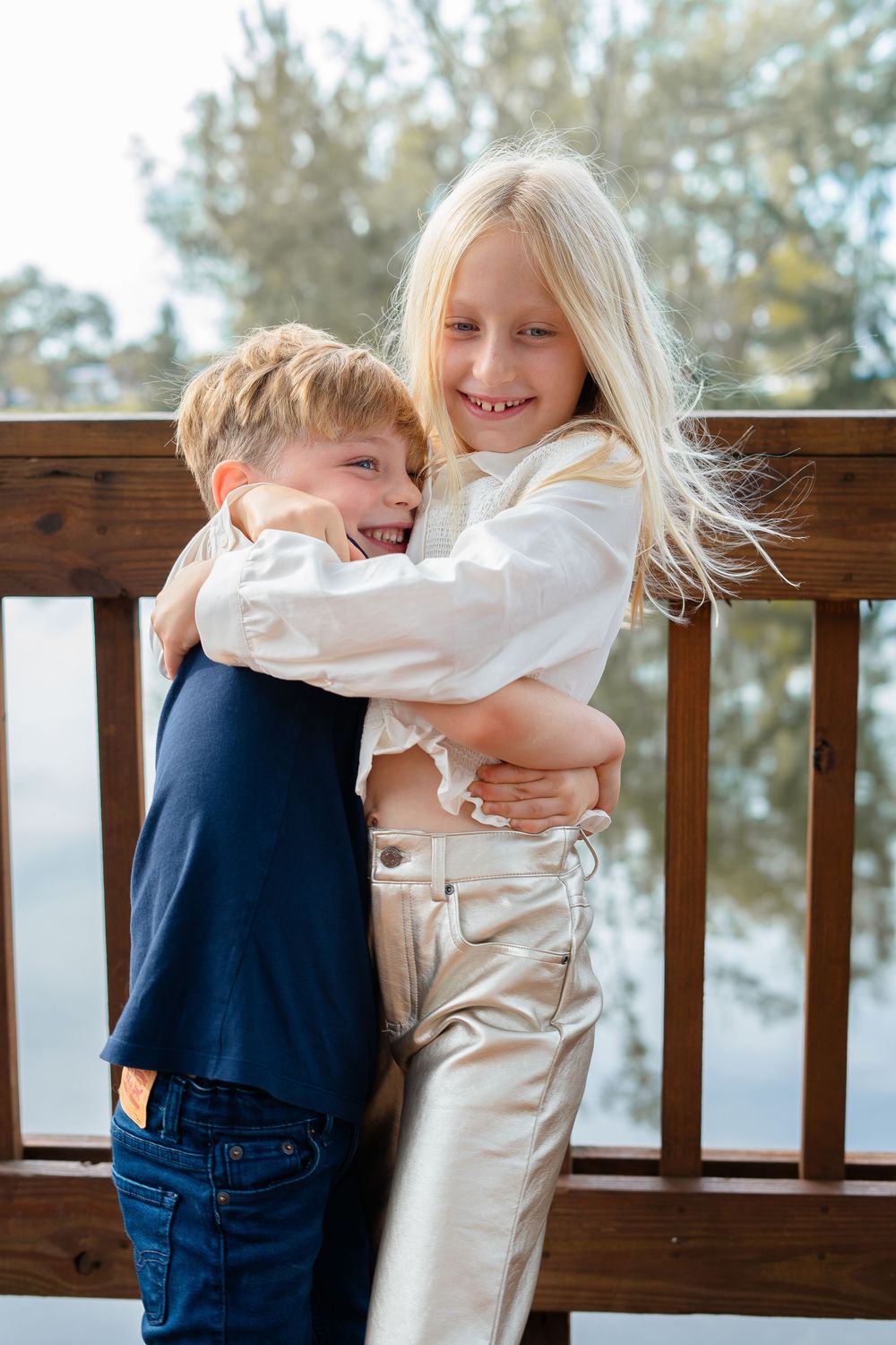 Two children share joyful laughter while playing together on a wooden deck railing with trees in the background.