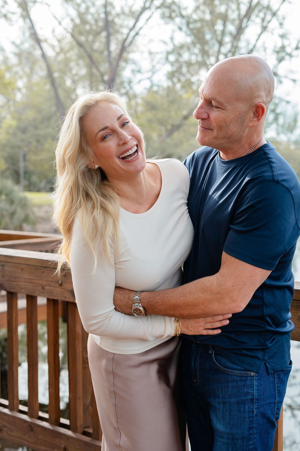 A couple shares joyful laughter while embracing on a wooden deck with trees visible in the background.