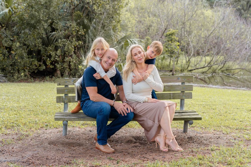 A family enjoys a tender embrace while sitting together on a wooden bench in a sunlit outdoor setting.