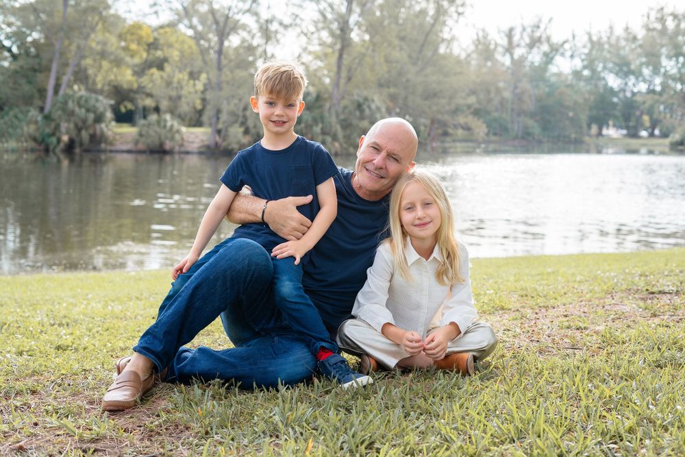 A candid outdoor family photo session on green grass with lake and trees in the background.