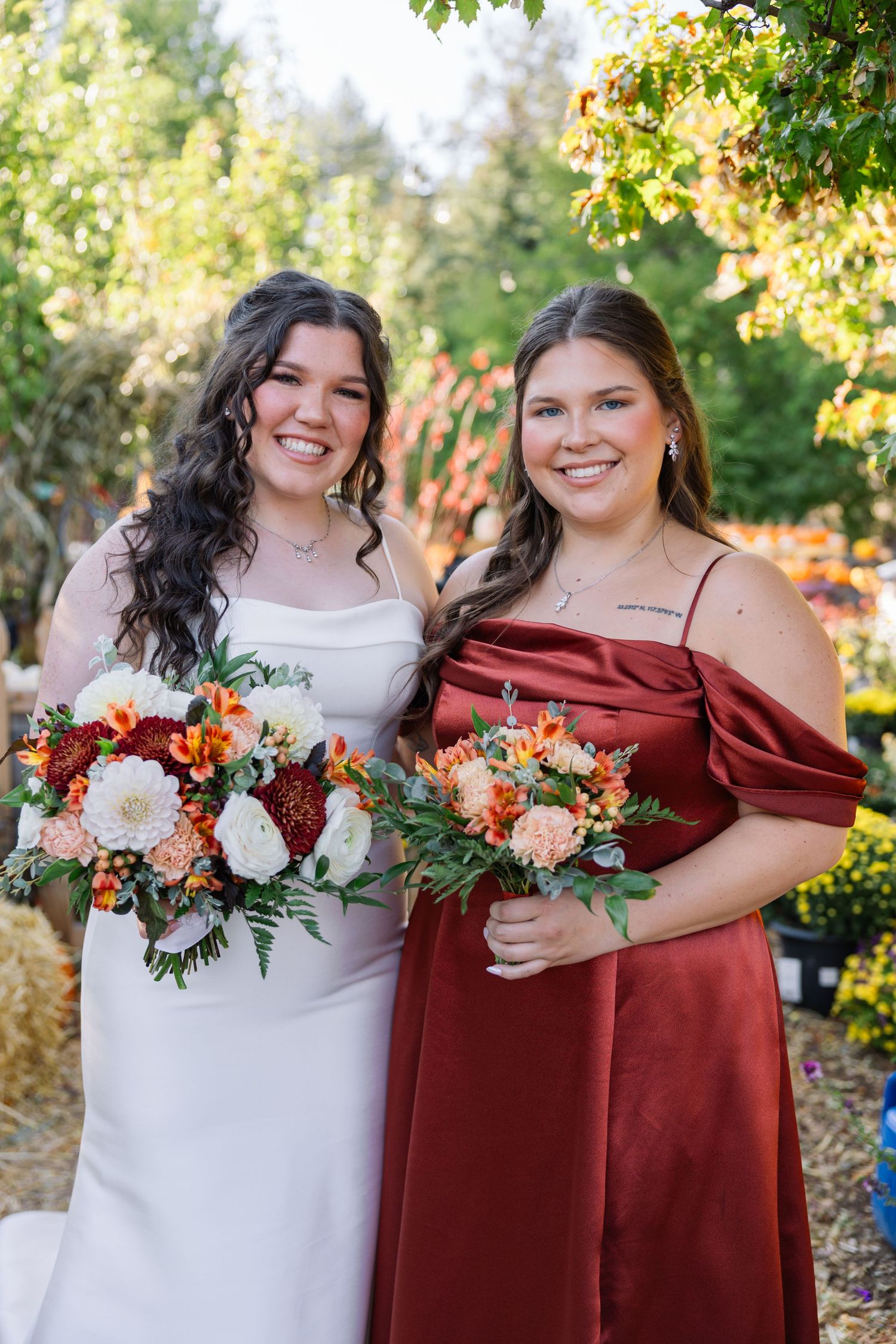 Two people in white and burgundy dresses holding autumn-inspired floral bouquets pose together outdoors on a sunny day.