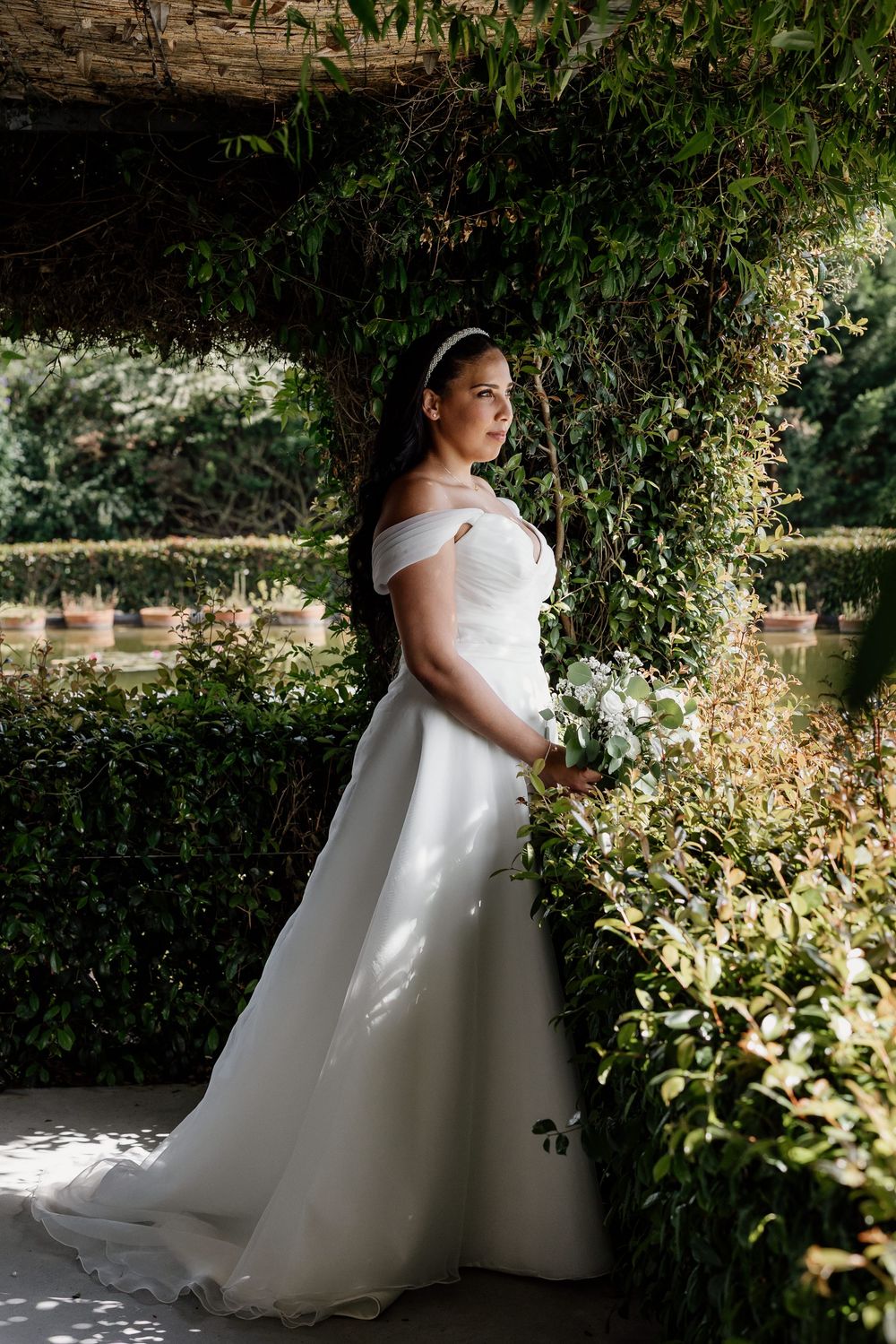 An elegant off-shoulder white wedding gown glows in dappled sunlight against a backdrop of lush garden greenery.