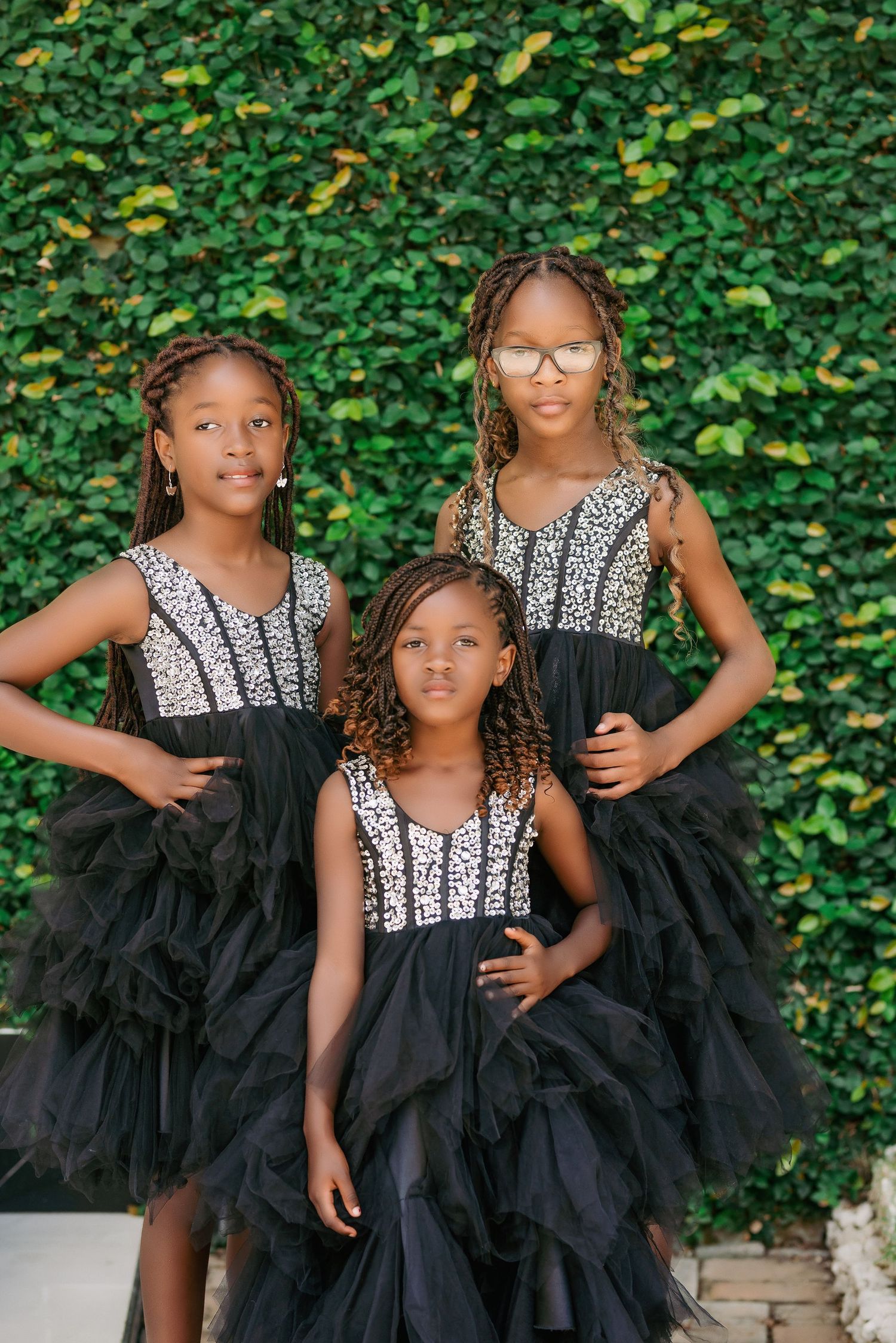 Three young children in matching black and white formal dresses pose against a leafy green hedge background.