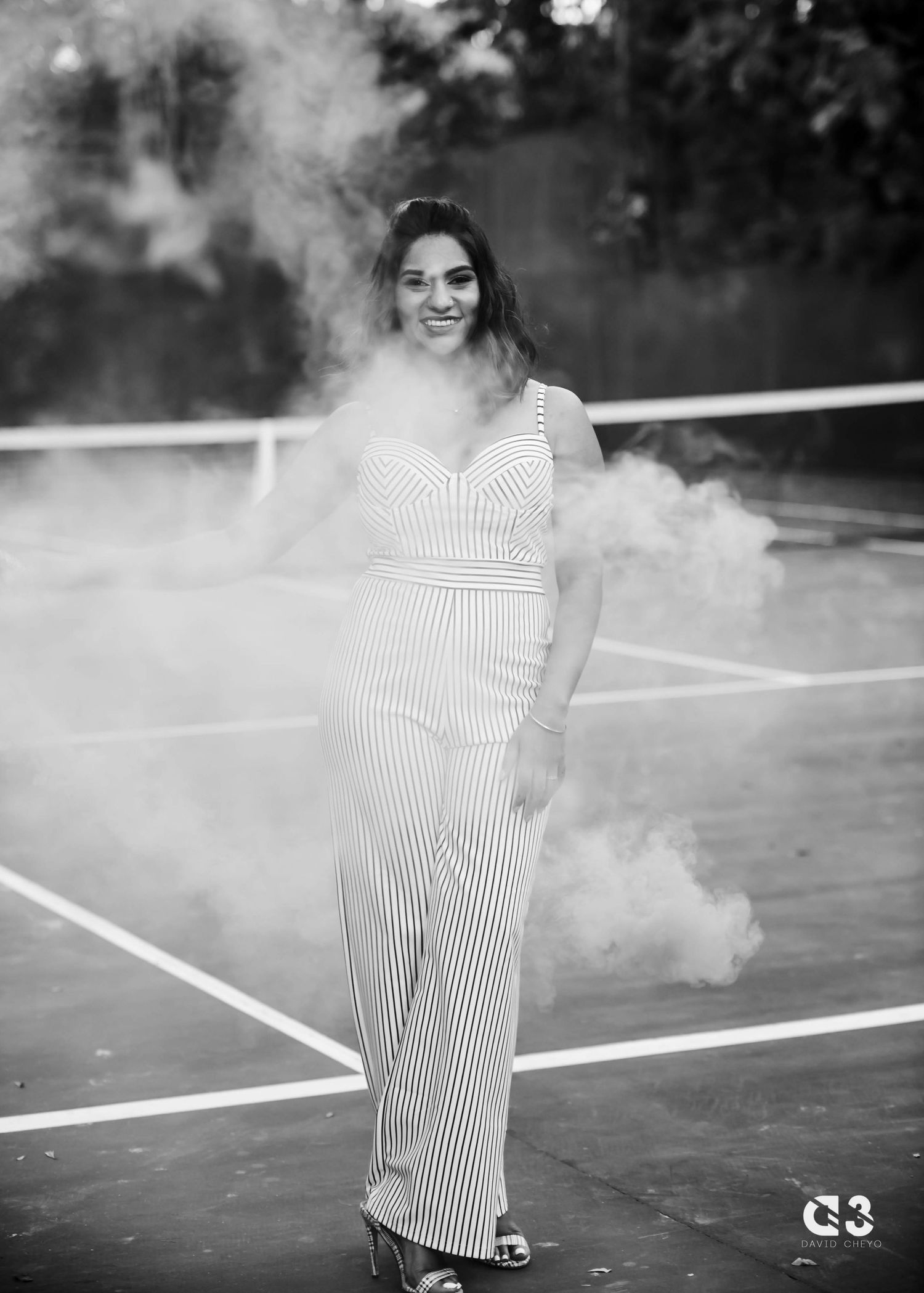 A woman poses on a tennis court in a striped jumpsuit while surrounded by white smoke effects.