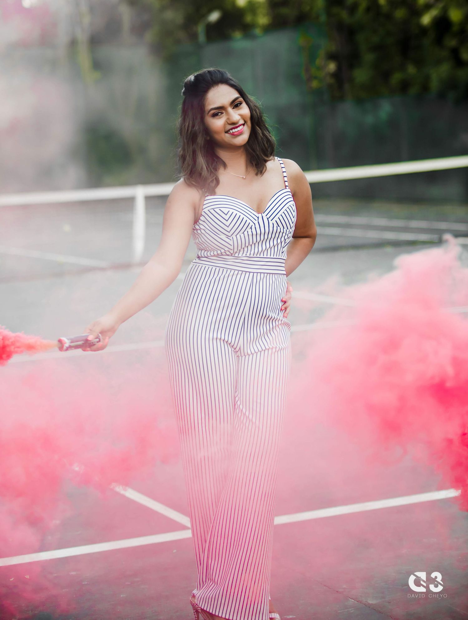 A woman in a striped jumpsuit poses with pink smoke effects on a tennis court.