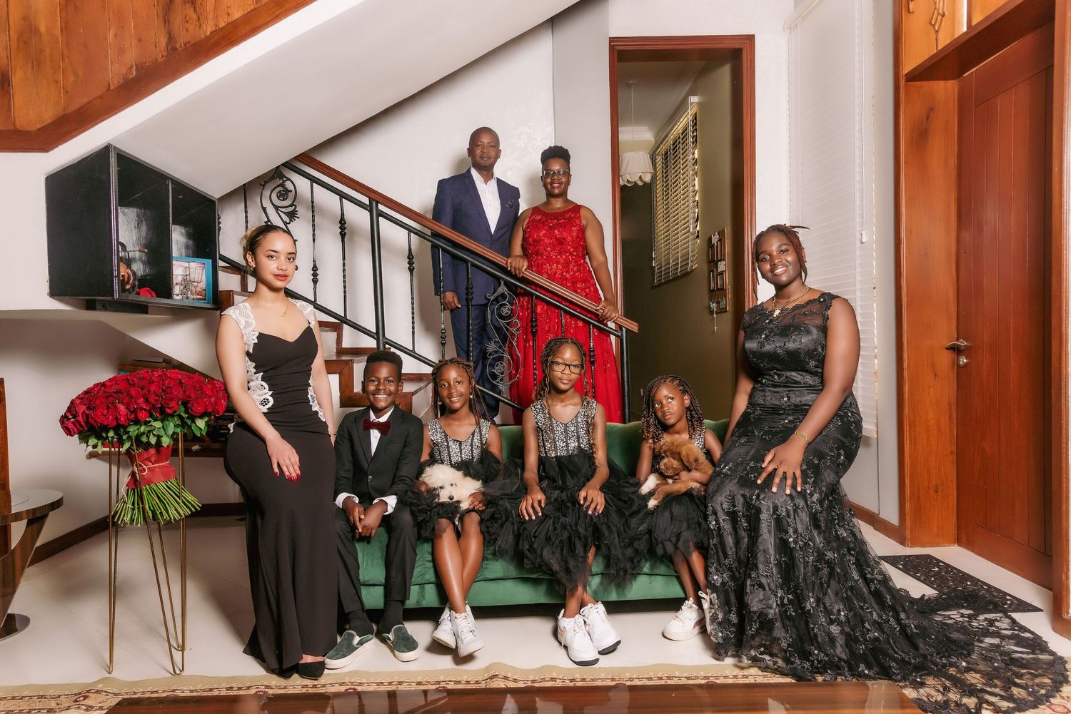 Formal family portrait of elegantly dressed people gathered on stairs with red roses in the foreground.