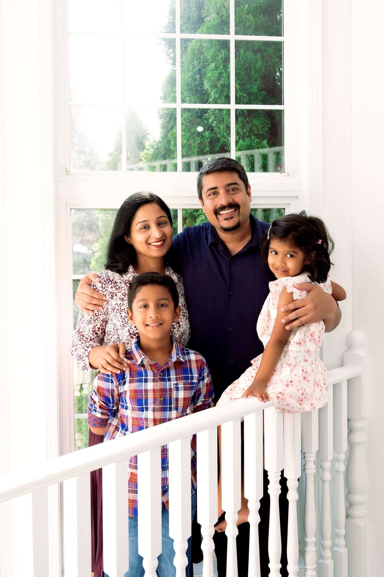 Family portrait of parents and two children standing by a white railing near a window.