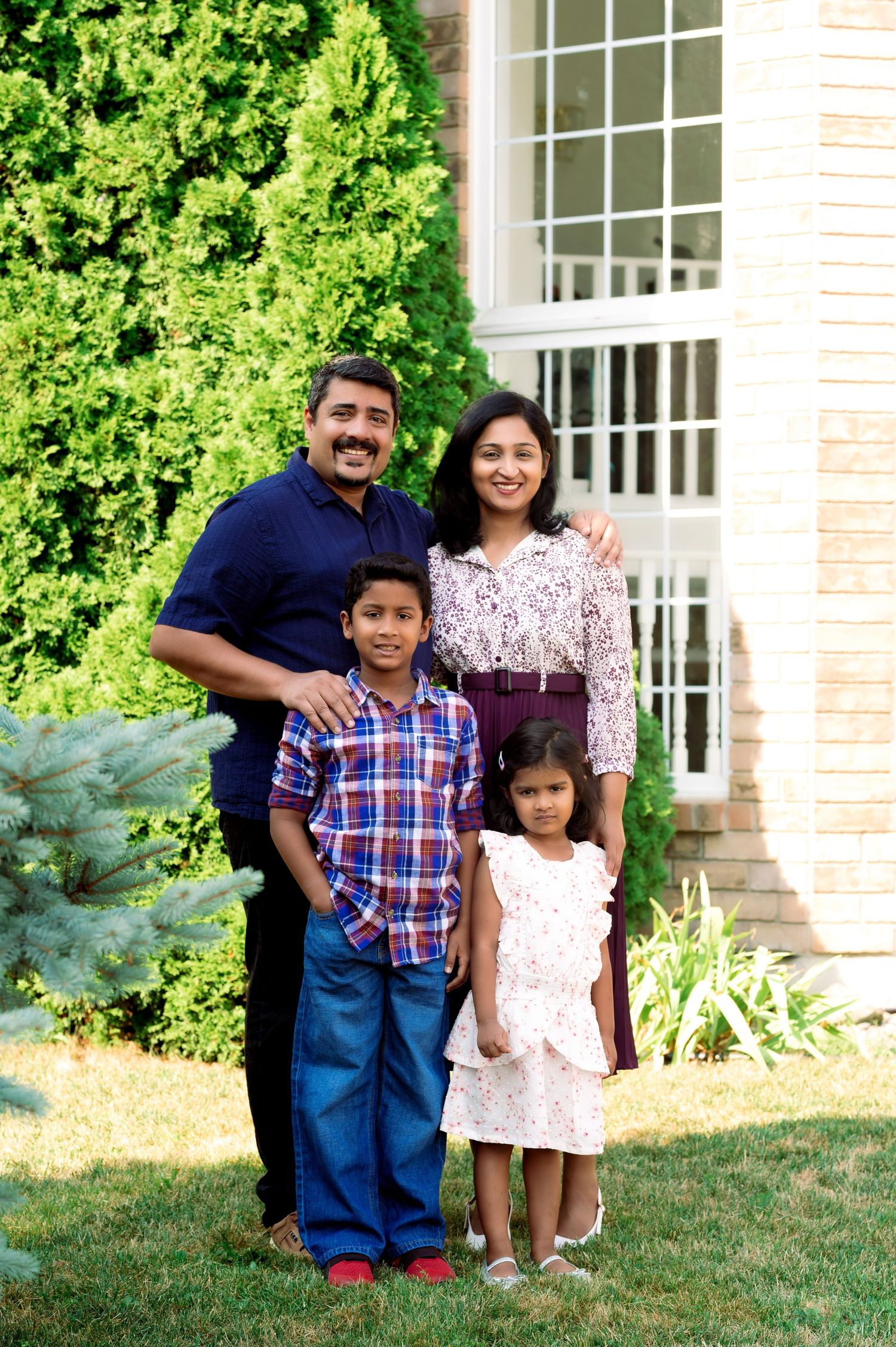 A family of four stands together outdoors in front of lush green bushes and a white house.