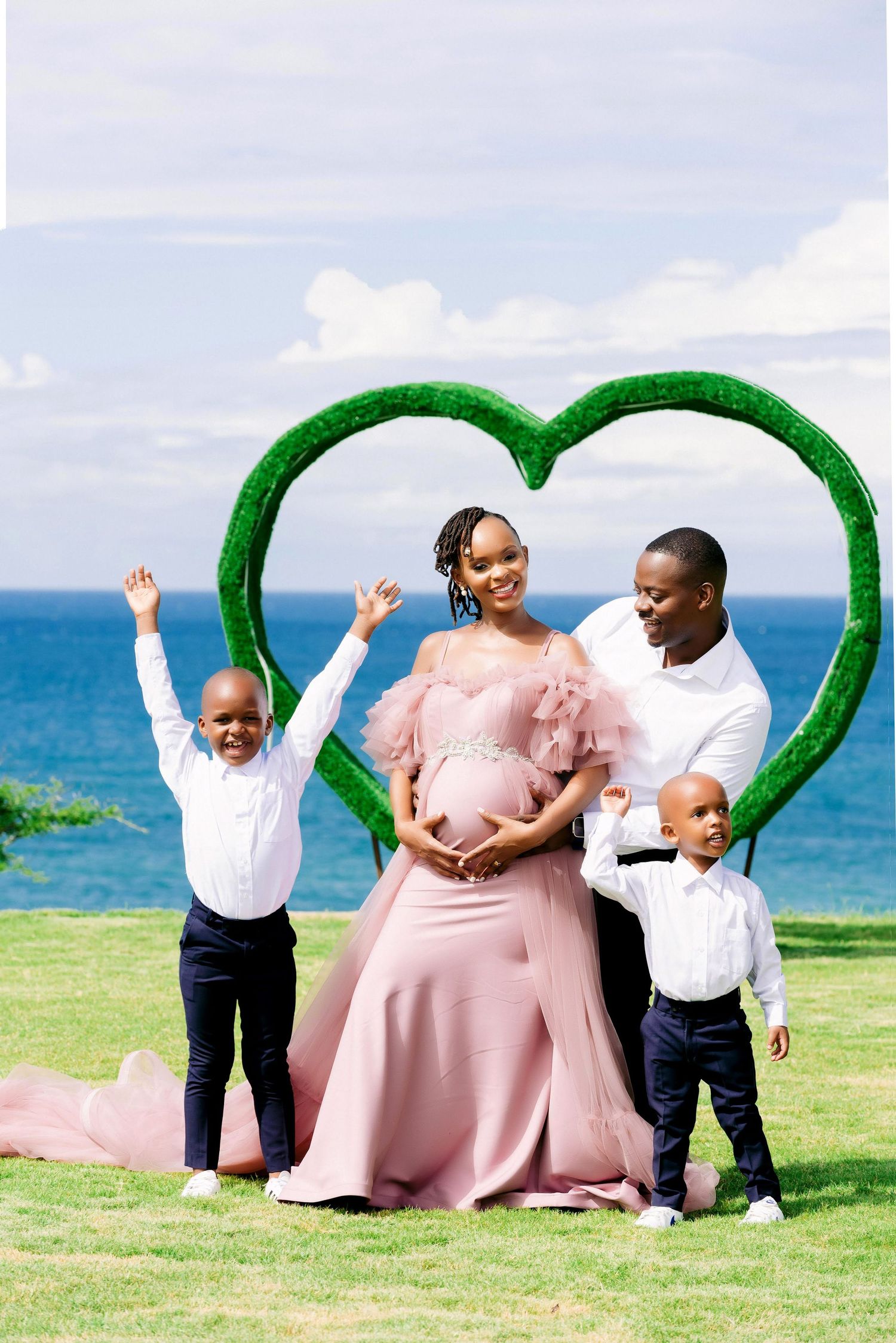 A family poses inside a green heart frame by the ocean with two children jumping joyfully.