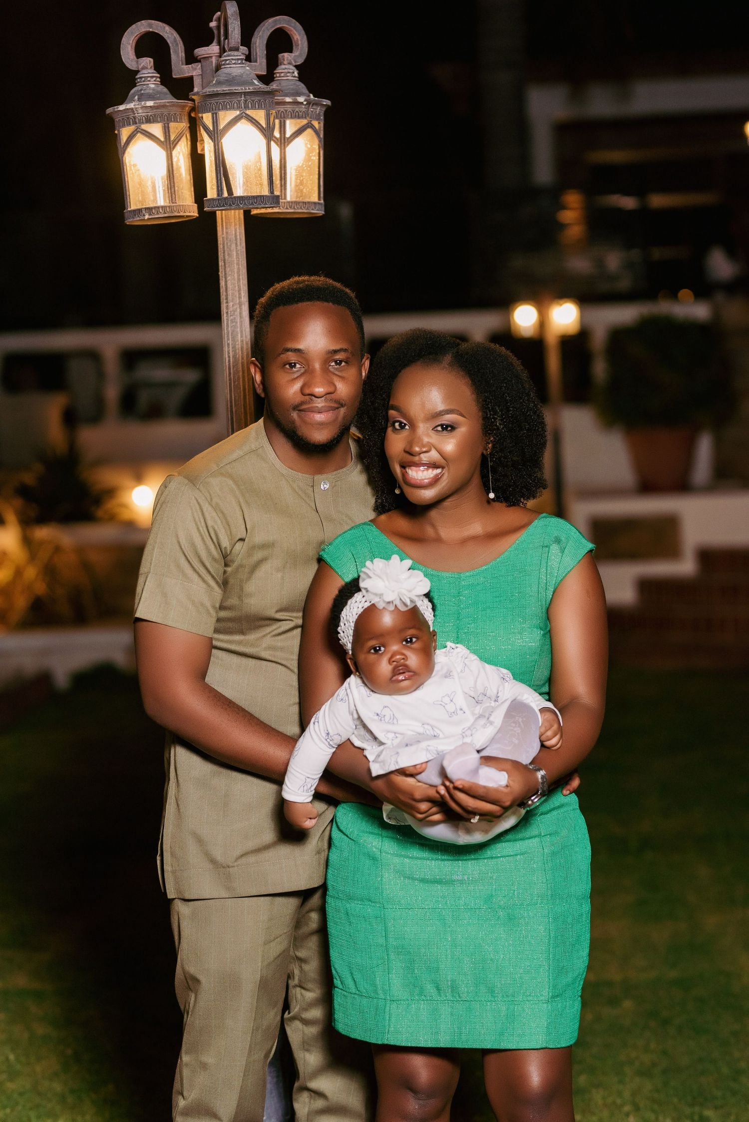 Family portrait at night under decorative lamppost with person in green dress holding baby in white.