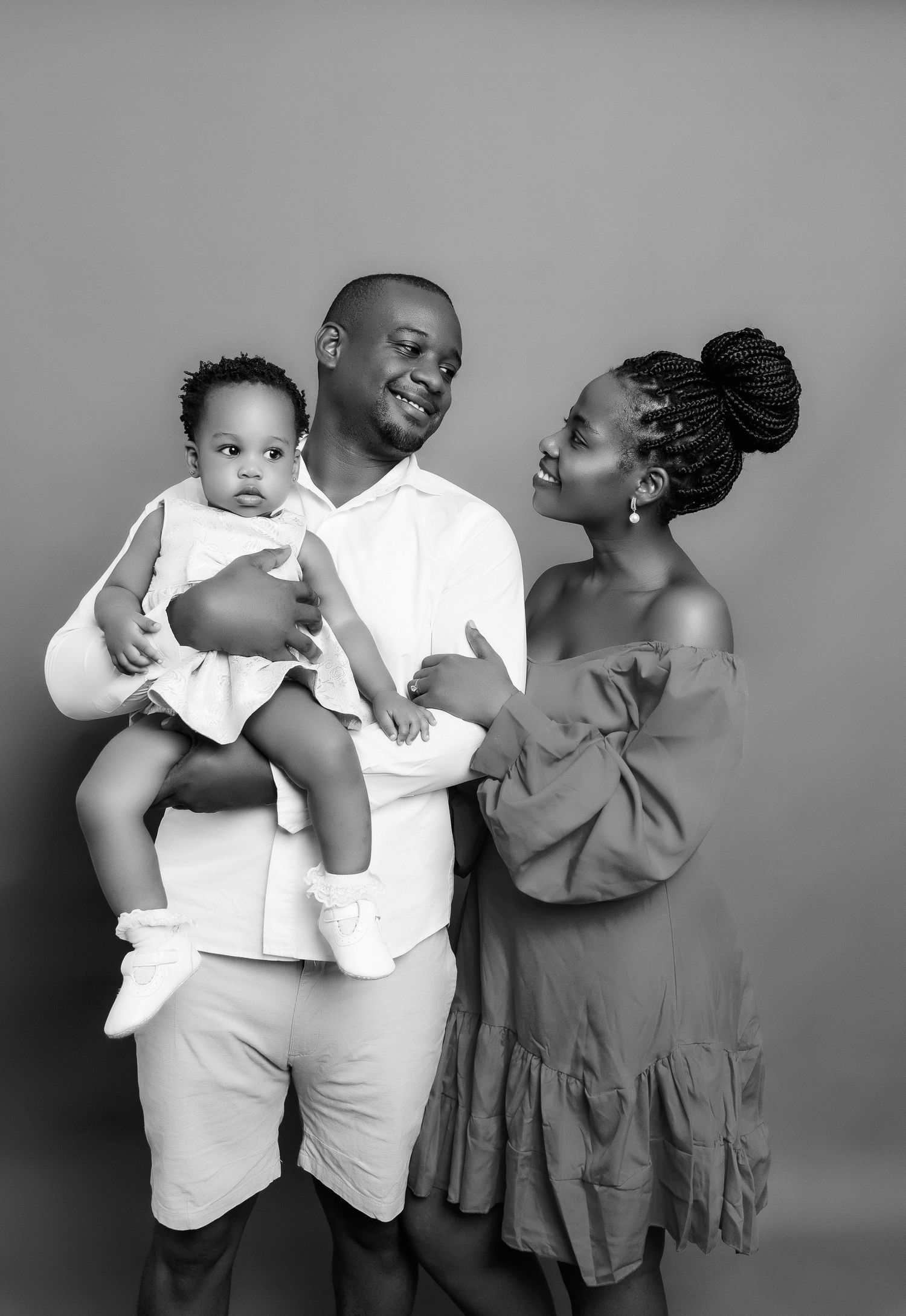 A family of three poses in casual white outfits against a grey background for a portrait.