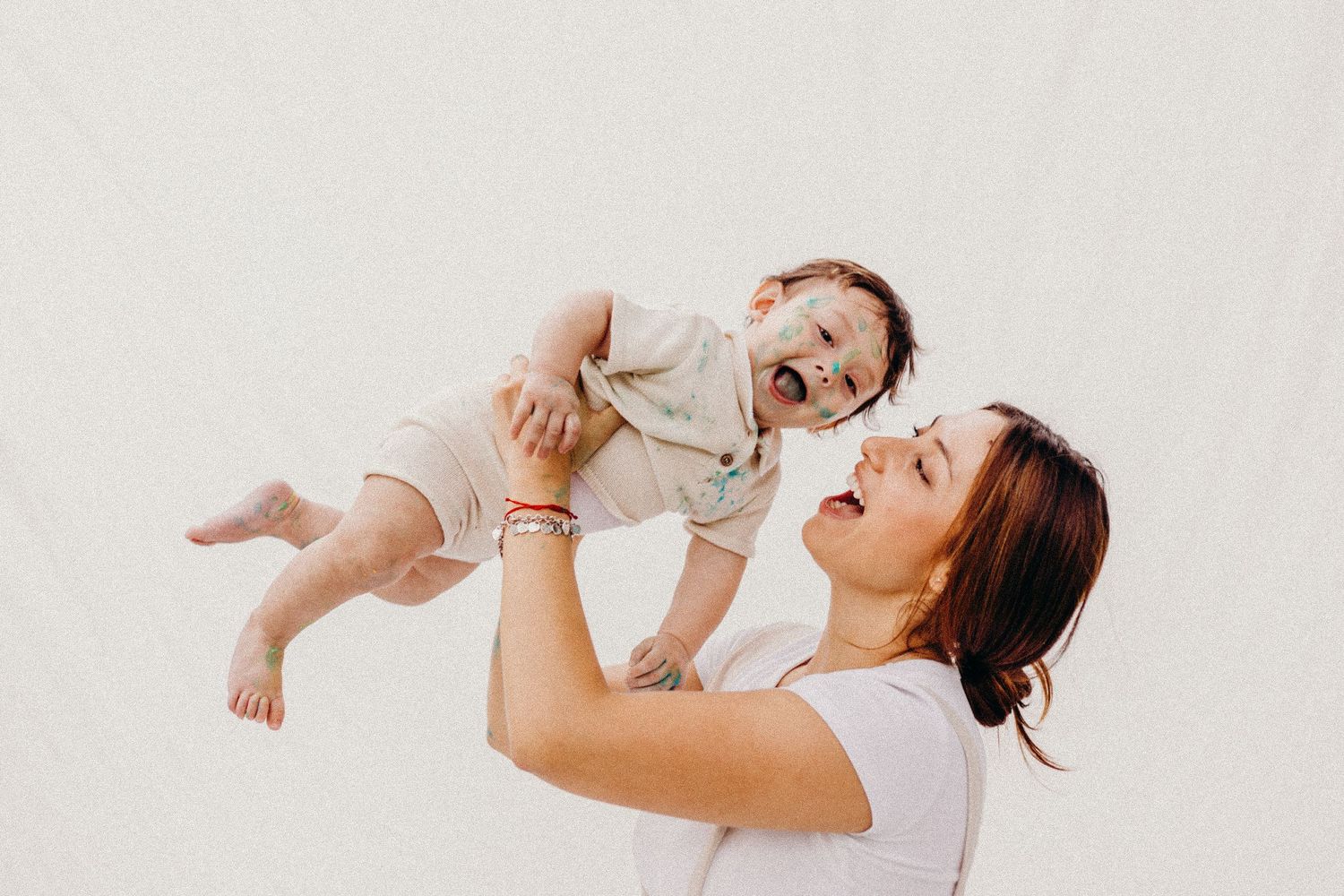Parent lifts laughing baby in white onesie against neutral background in candid moment.