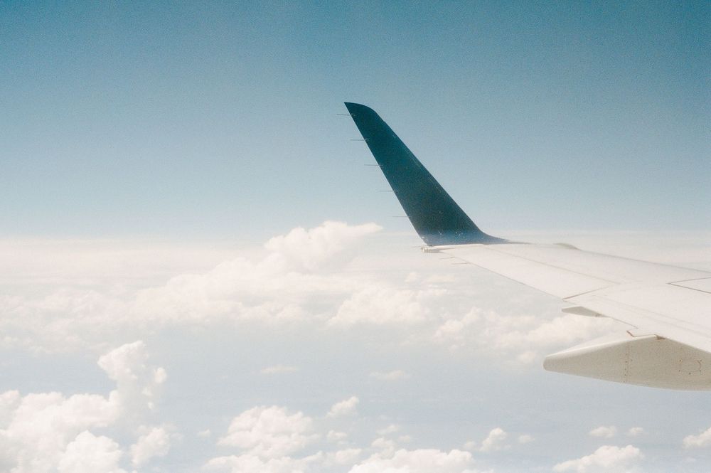 Airplane wing cutting through fluffy white clouds against a bright blue sky on a clear day.