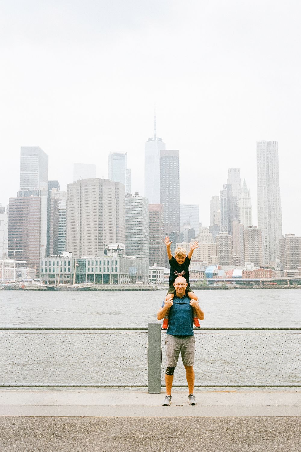 People enjoying a playful moment along the waterfront with the New York City skyline rising behind them in misty weather.