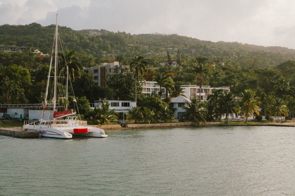Panoramic view of a tropical marina along the coastline with sailboats anchored in calm blue waters and palm trees onshore.