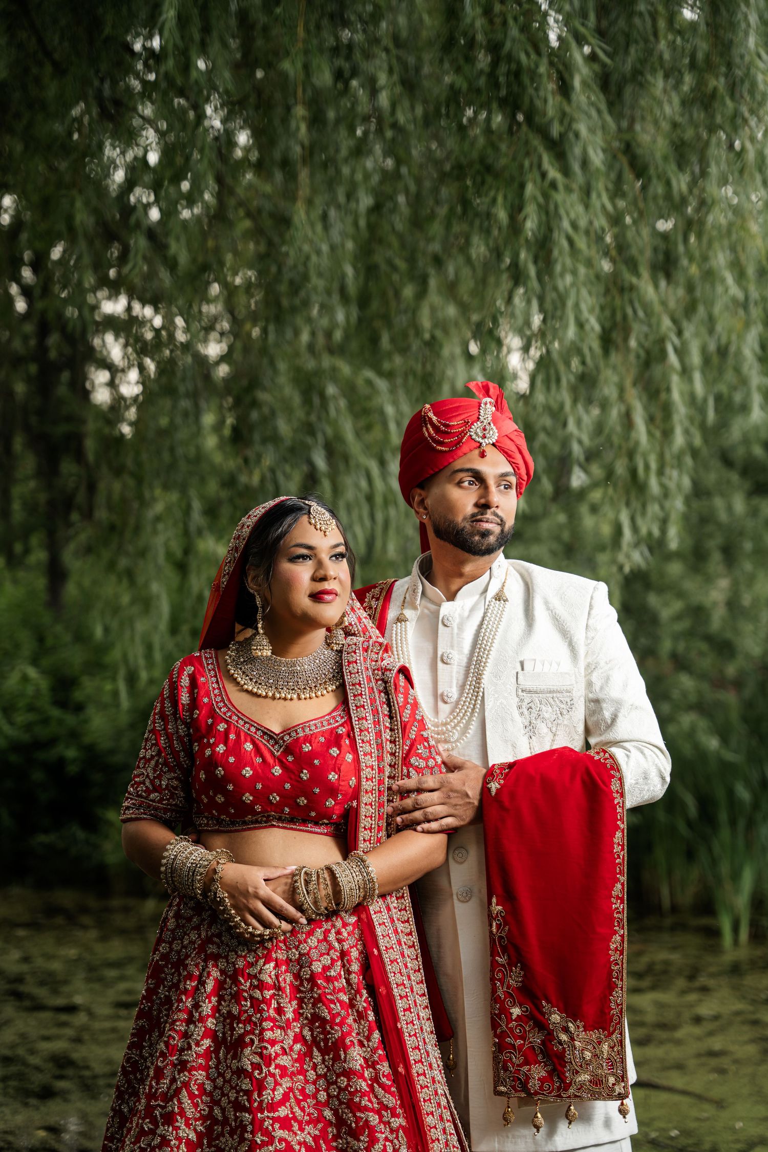 Traditional Indian wedding couple poses beneath willow trees in red and white formal attire.