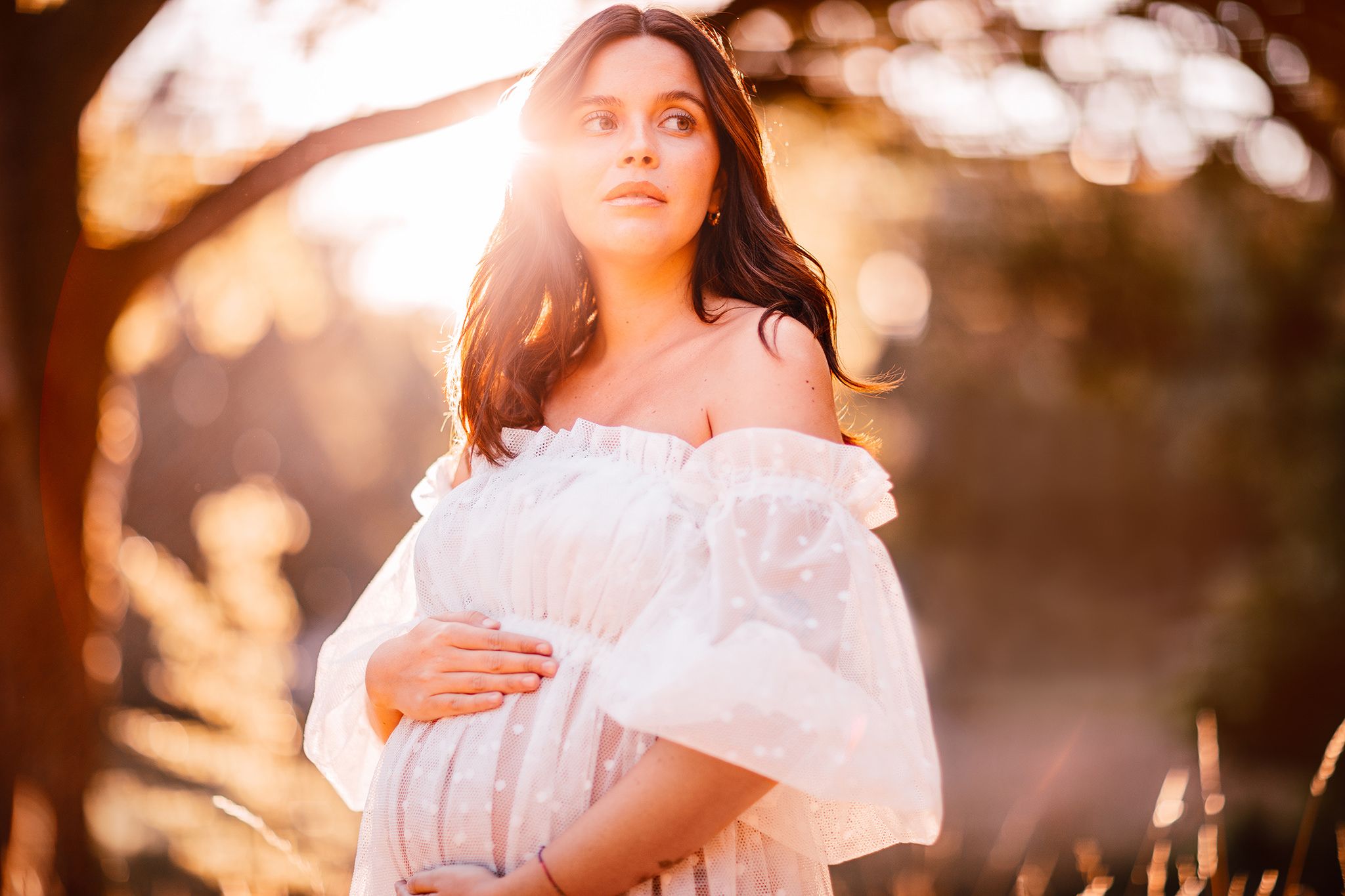 Person in a white dress poses outdoors during golden hour sunlight with a dreamy ethereal atmosphere.