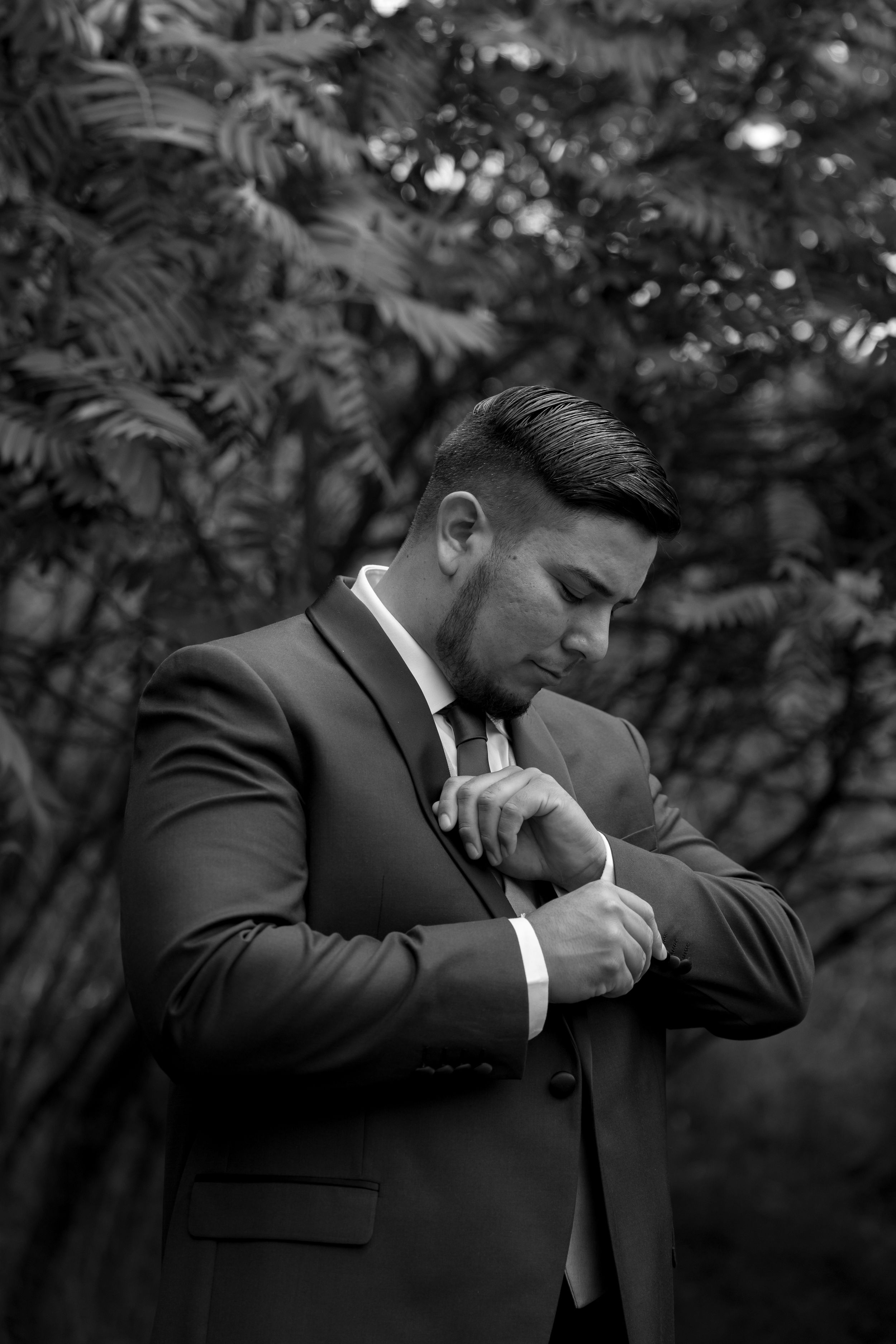 Black and white portrait of a person in formal suit adjusting their tie while standing outdoors with foliage in background.