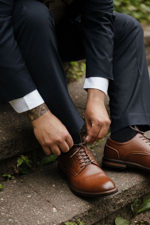 Close-up sequence showing hands tying brown leather dress shoes on stone steps with greenery in background.