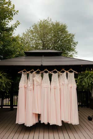 Row of blush pink bridesmaid dresses hanging on wooden hangers displayed outdoors on a wooden deck.