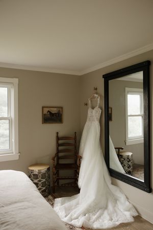 A wedding dress hangs elegantly in a sunlit room with white walls and a large mirror.