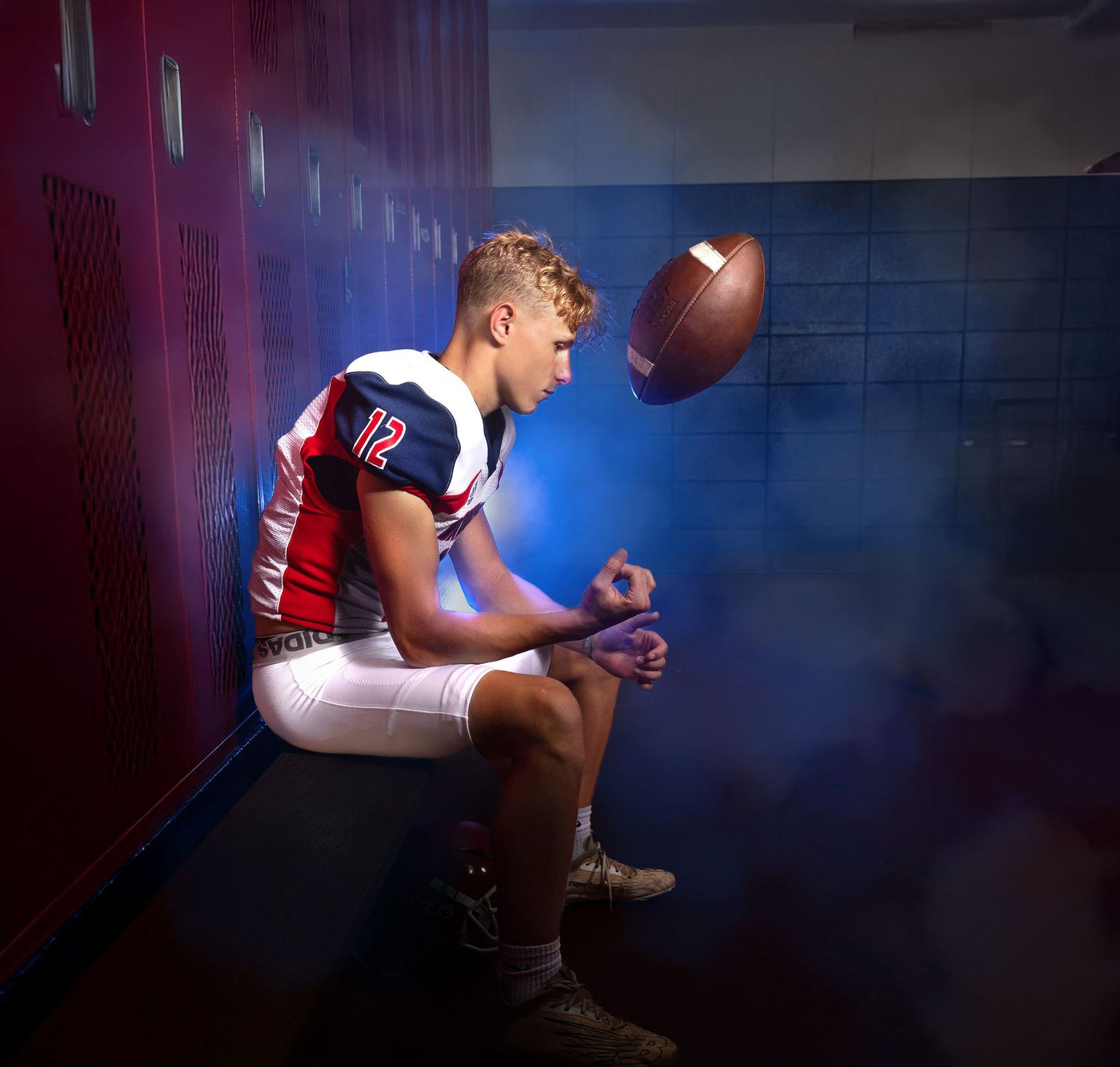 Football player sits in locker room hallway flipping a football into the air in dramatic lighting.