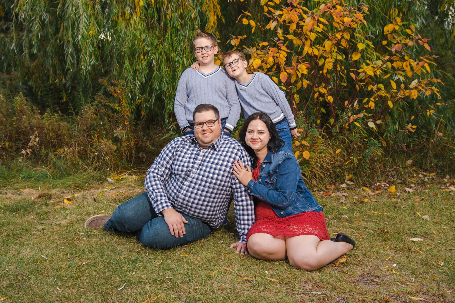 A family poses together outdoors on grass with autumn foliage and willow trees in the background.