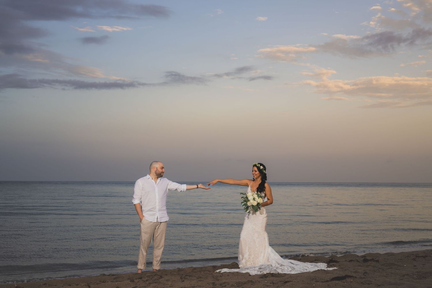 A bride and groom hold hands while standing apart on a beach during their sunset wedding photoshoot.