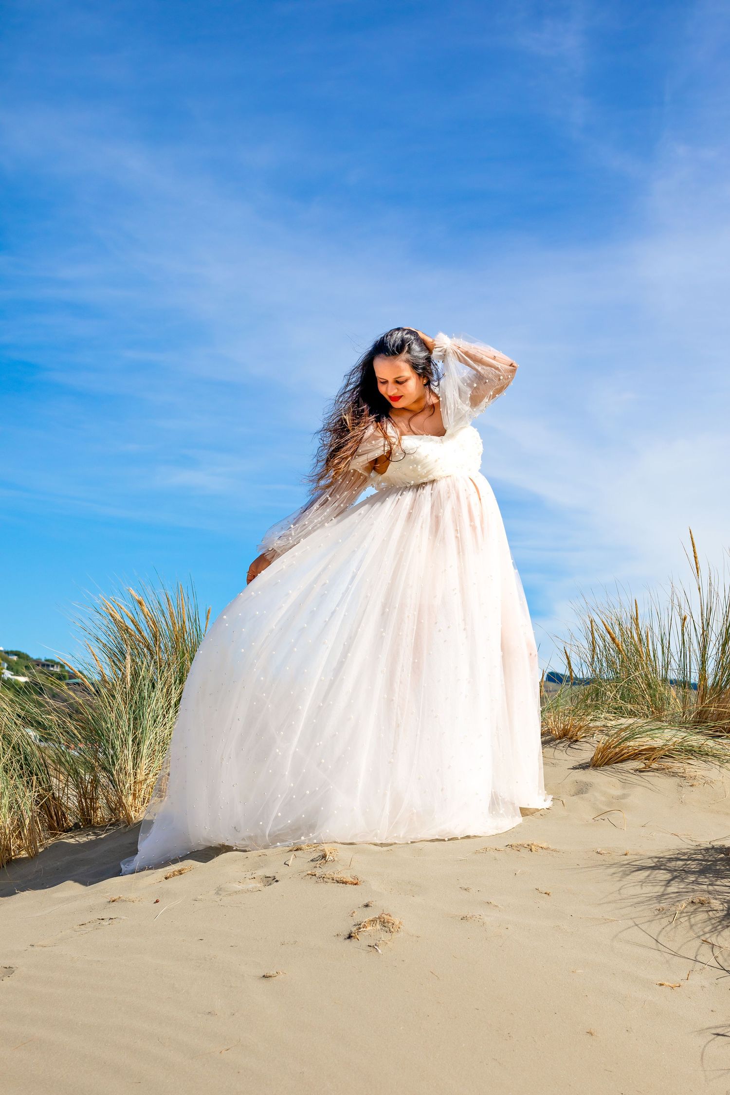 Flowing white dress caught in the wind on a sandy beach dune with tall grass and bright blue sky in the background, taken at Castlepoint.