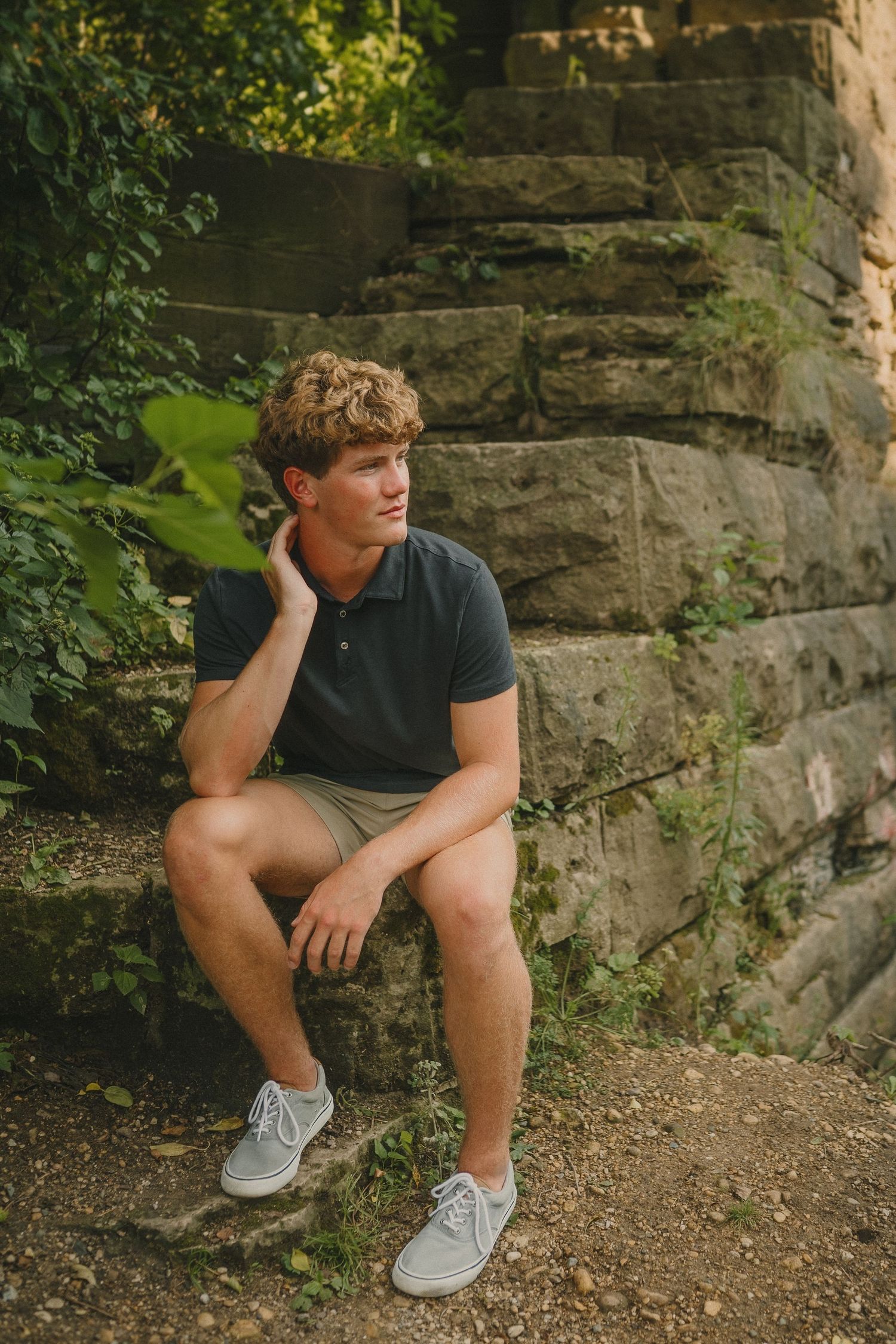 Person in casual attire sits thoughtfully on stone steps surrounded by greenery and natural lighting.