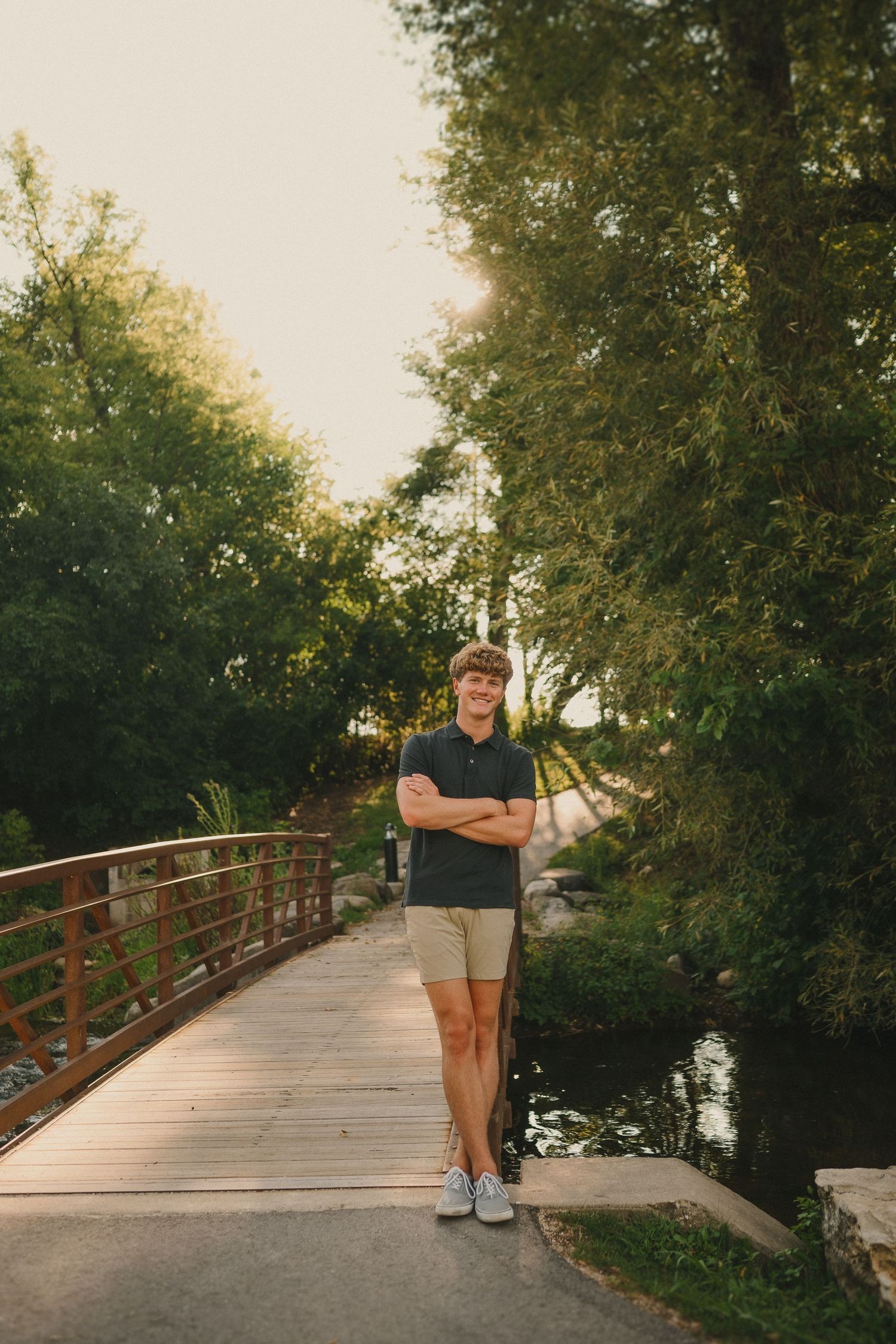 Person stands on wooden bridge in park wearing navy polo and khaki shorts on summer day.