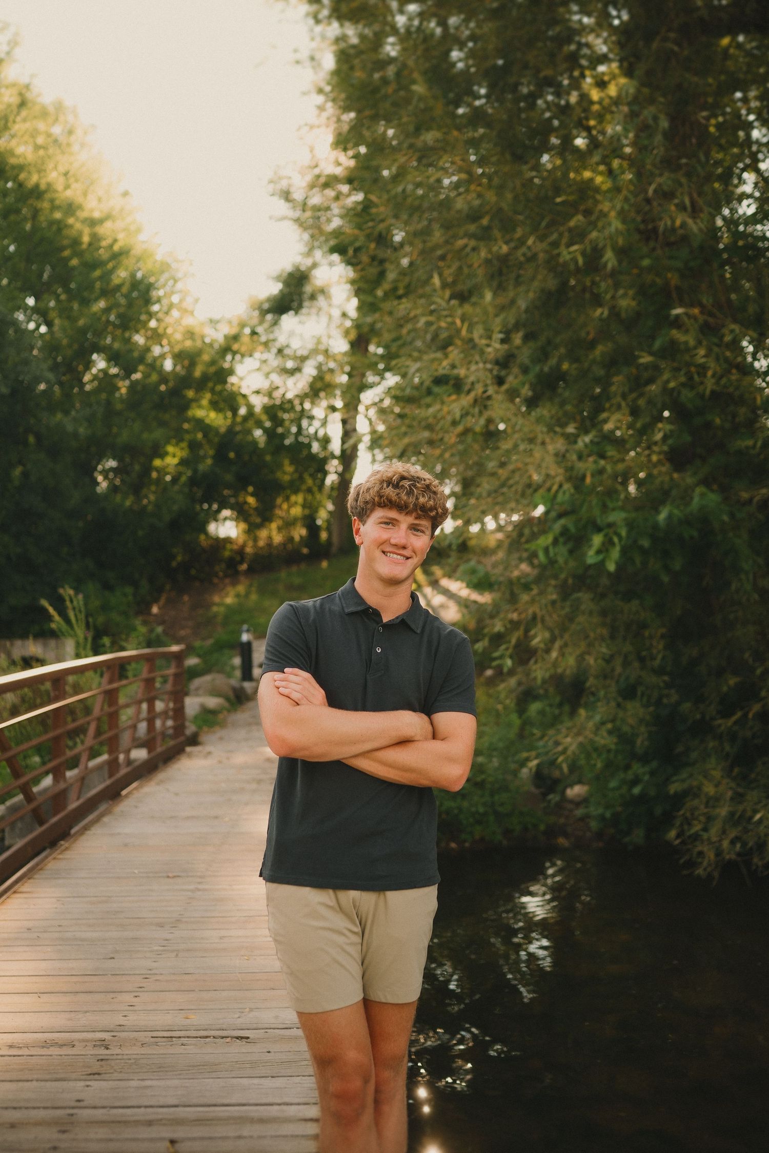 A person poses with crossed arms on a wooden bridge pathway surrounded by lush green trees in warm afternoon light.