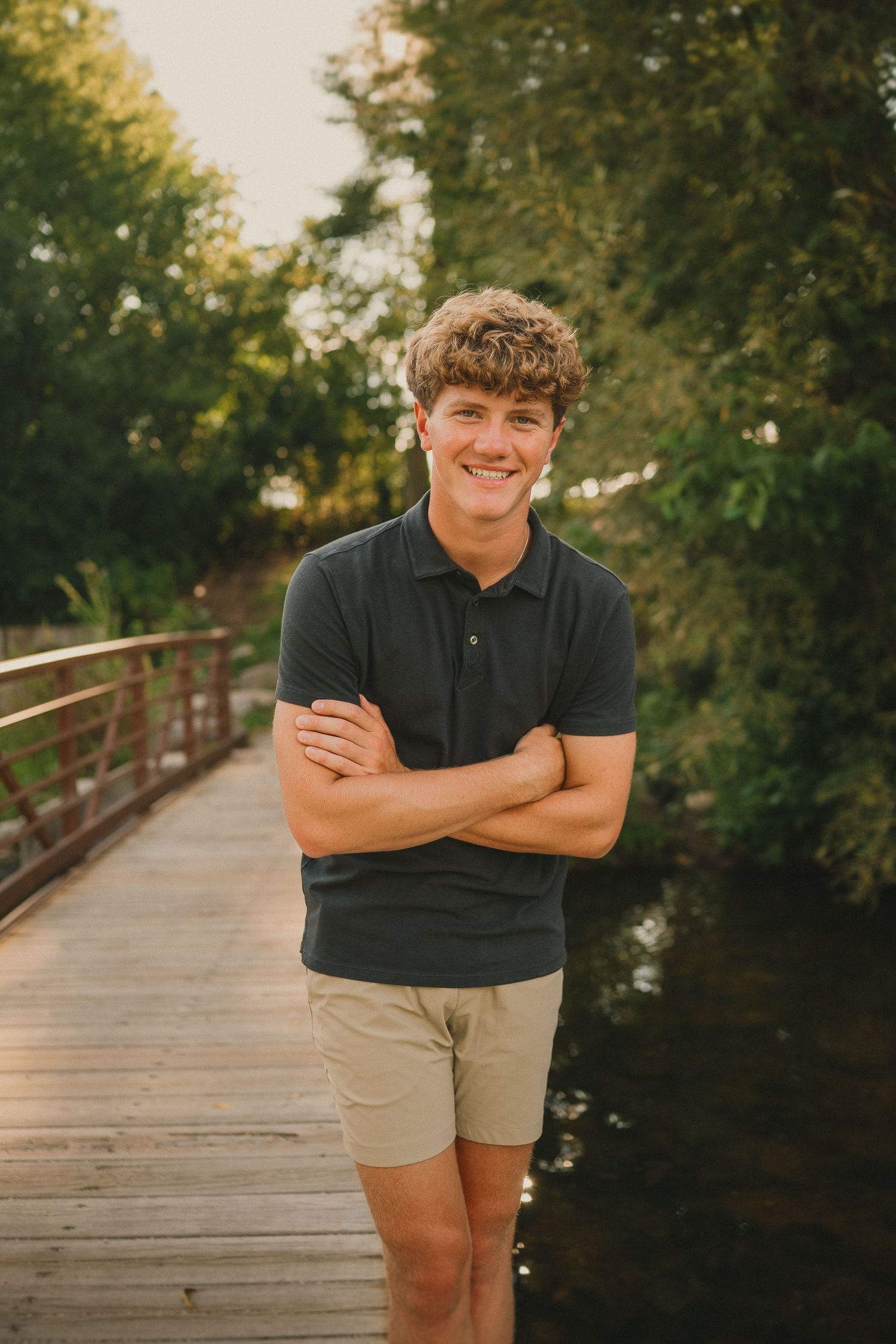 Person in a black polo shirt stands smiling on a wooden bridge walkway at sunset.