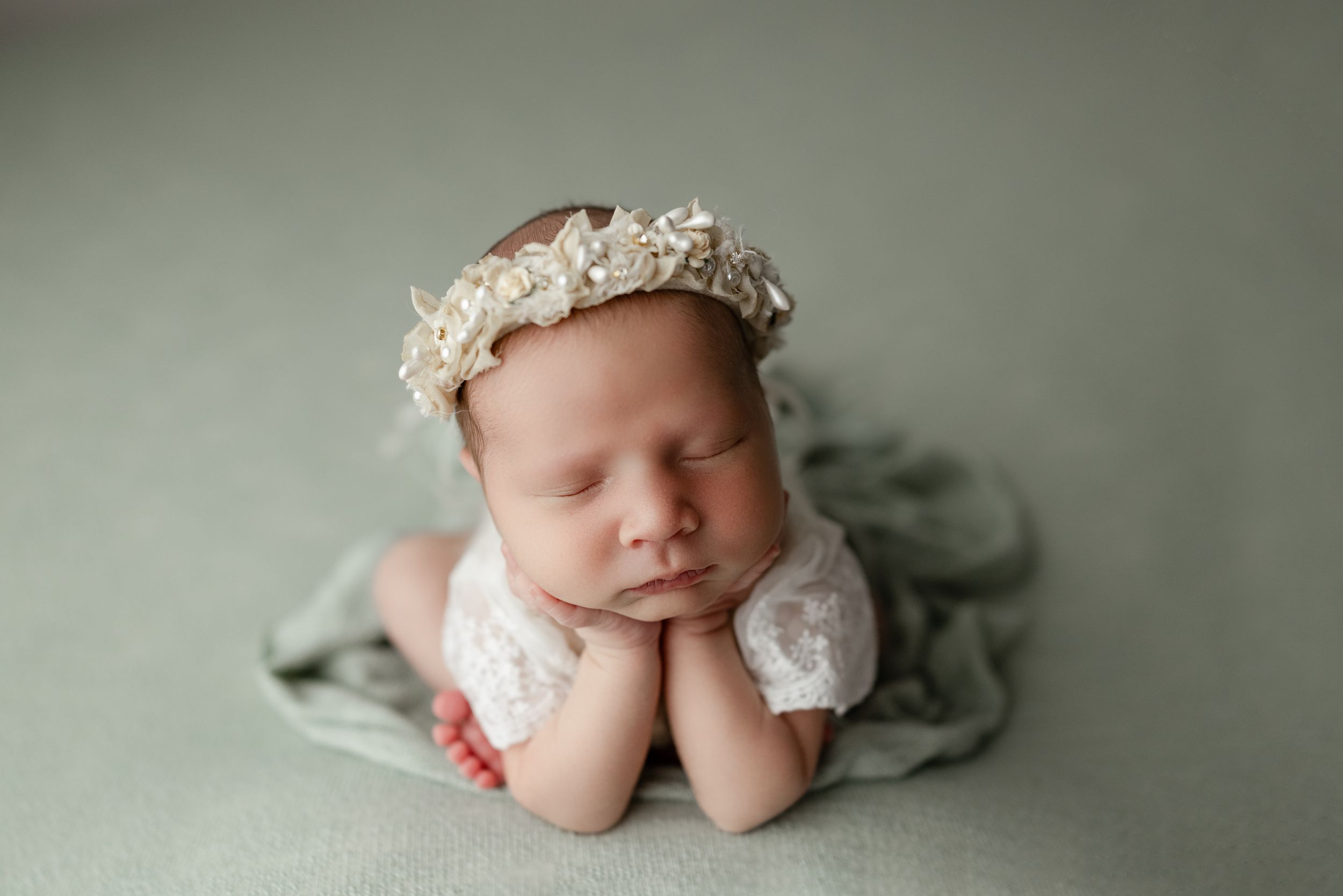 Sleeping newborn wearing lace headband rests head on hands while lying on soft sage green blanket.