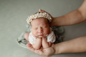 Sleeping newborn in white lace outfit and floral headband rests peacefully in parent's hands on grey backdrop.