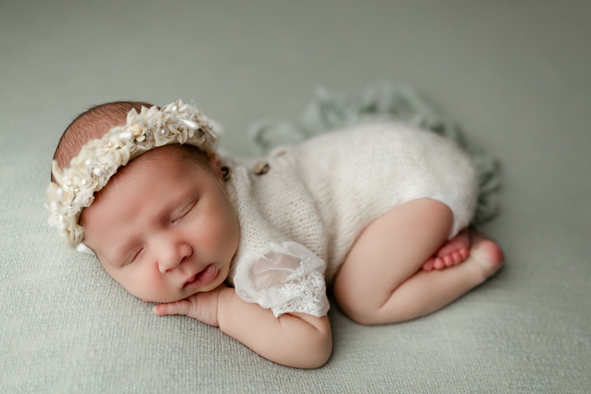 A sleeping newborn in a white lace outfit and delicate headband rests peacefully on a mint green backdrop.