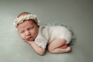 Sleeping newborn dressed in soft white romper and floral headband rests peacefully on mint green backdrop.