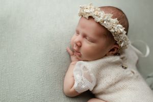 Sleeping baby wearing a delicate floral headband and lace outfit lying on a soft gray background.