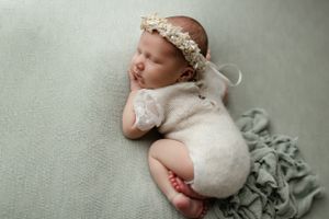 Sleeping newborn in white lace wrap and floral crown rests peacefully on soft sage green blanket.