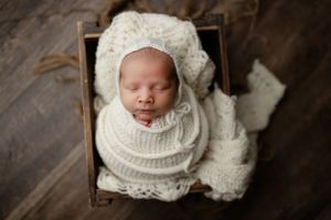 Sleeping newborn wrapped in cream knitted blankets nestled in wooden box on hardwood floor.
