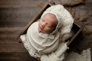 Sleeping newborn wrapped in cozy white knit blankets rests peacefully in rustic wooden box setting.