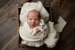 A sleeping newborn swaddled in cream-colored knit blankets holds a small stuffed animal while lying in a wooden box.