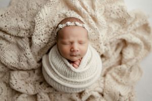 Peaceful newborn swaddled in cream knit wrap rests on delicate lace fabric backdrop.