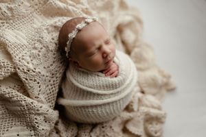 A sleeping newborn baby wrapped in a cream-colored knit blanket on soft lace fabric with a delicate headband.