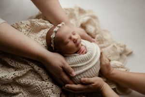A sleeping newborn swaddled in a white knit wrap is gently cradled by caring hands on lace fabric.