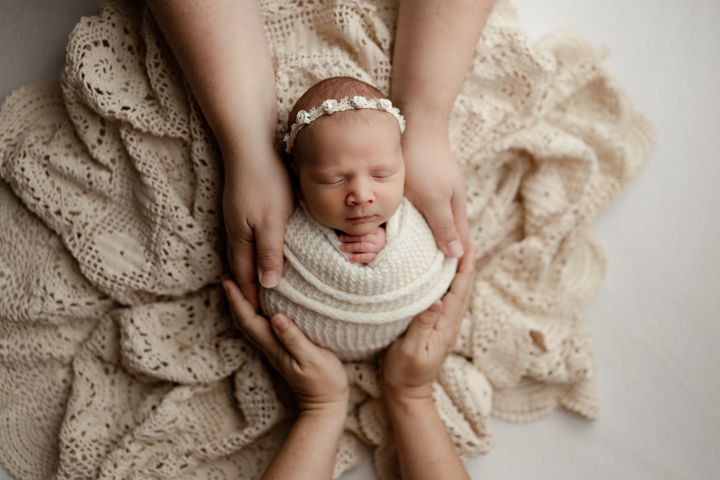 Overhead view of newborn baby peacefully sleeping in white wrap on beige lace blanket with parent's hands gently cradling.