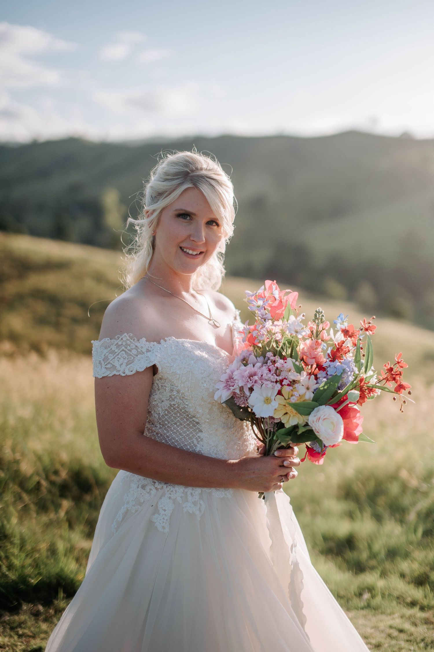 Bride holding vibrant bouquet poses in white gown against mountainous landscape.