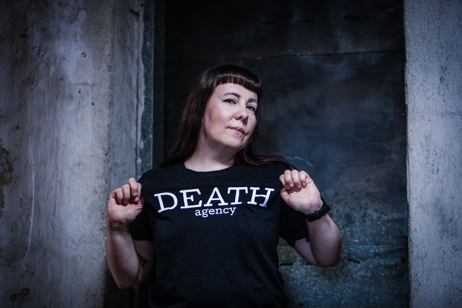 A person with bangs wearing a black t-shirt with DEATH text stands against a dark concrete wall.