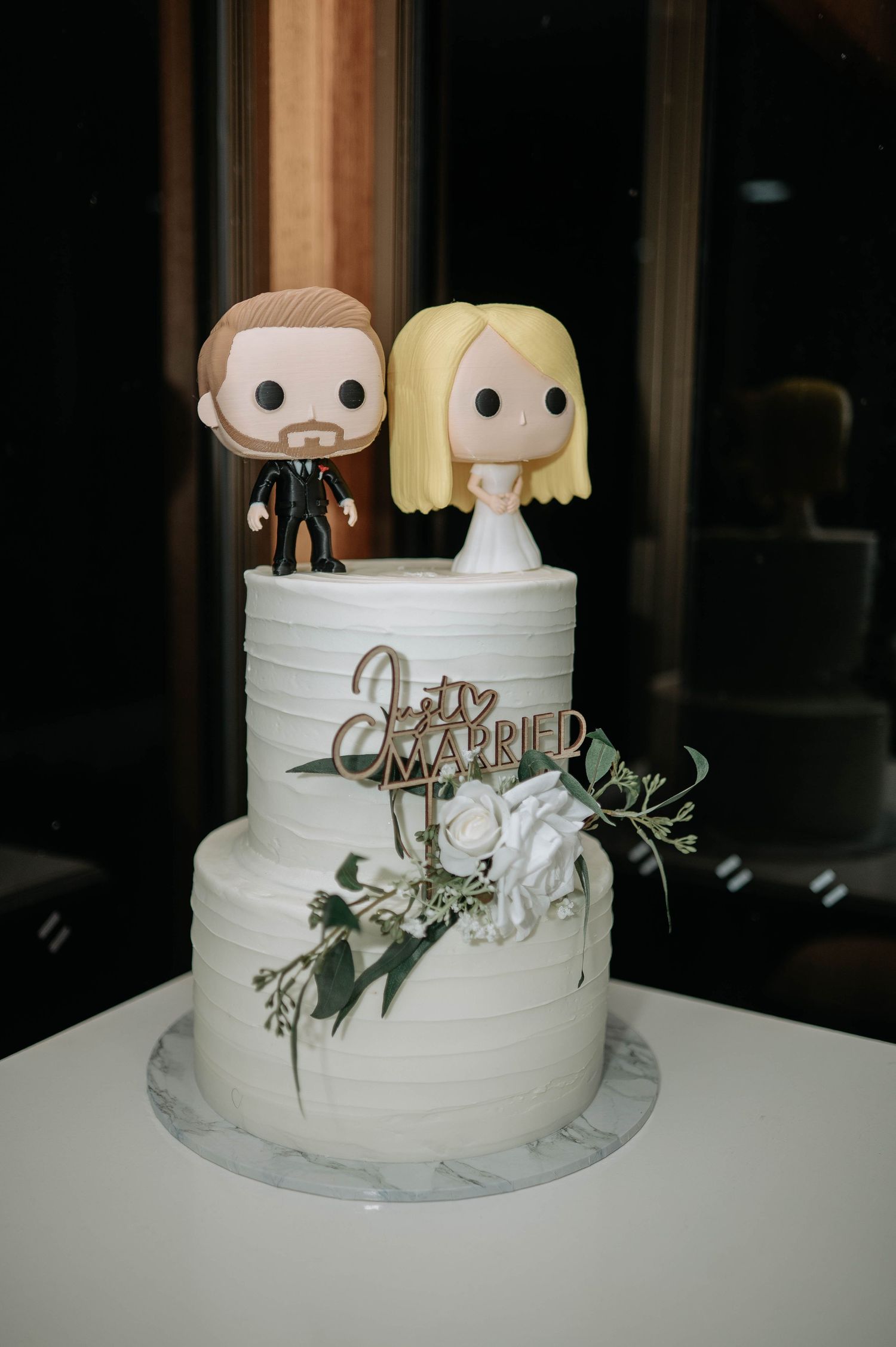 White wedding cake decorated with Funko Pop-style bride and groom toppers and greenery.