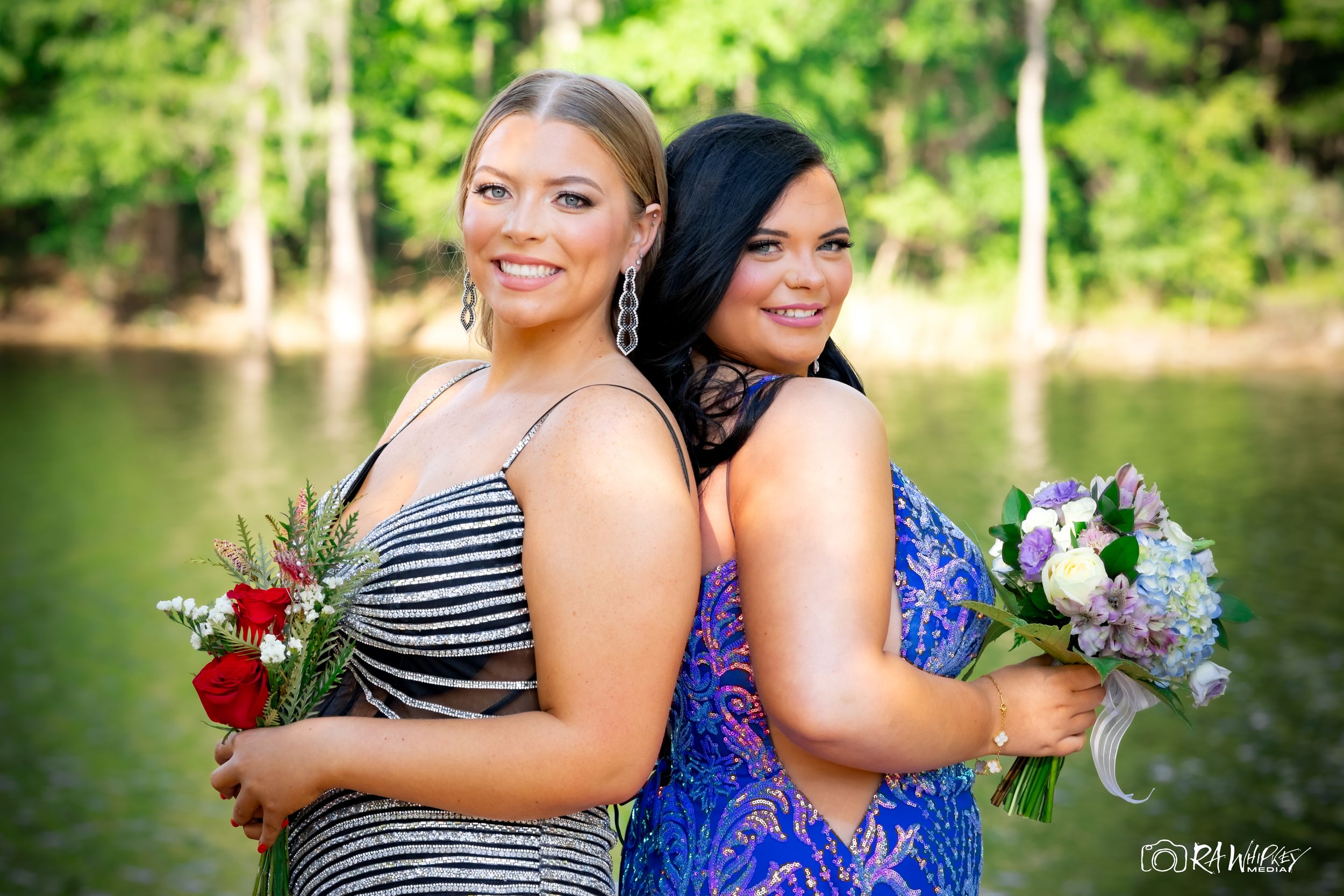Two women in formal dresses posing back-to-back with flower bouquets by lake.